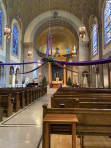 St. Joseph’s Memorial Chapel, featuring wooden pews, an altar with Christmas trees and white statues on either side, and a hanging Advent wreath as the center of the image.