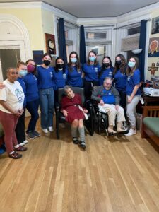 Nine girls, all wearing the same blue shirt, posing with three other older individuals, two of which are seated one in a wheelchair) and the third on the far left.